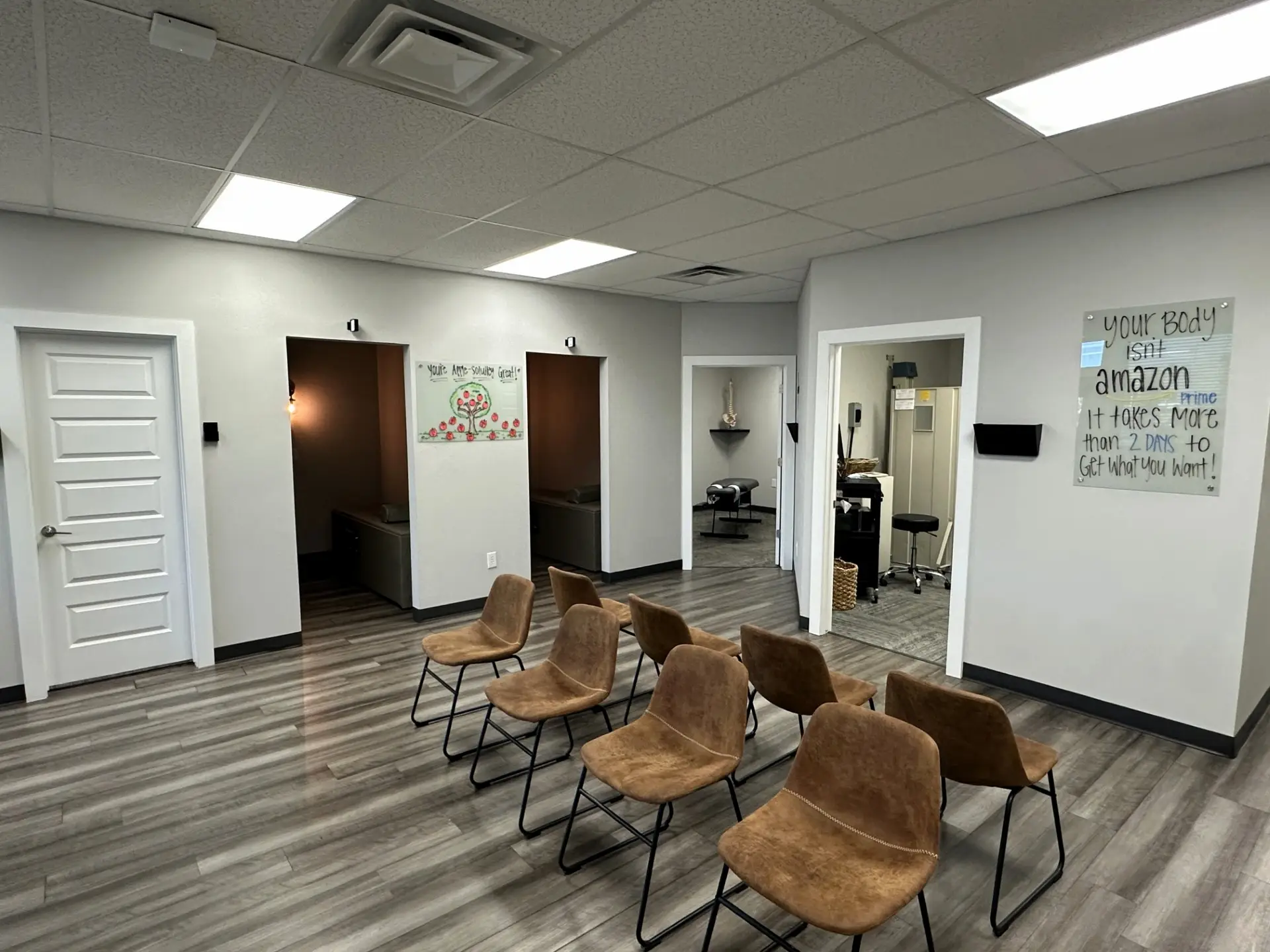 An empty waiting room with brown chairs, grey walls, and wood-look flooring. Motivational signs are visible, including one about the body not being Amazon Prime.