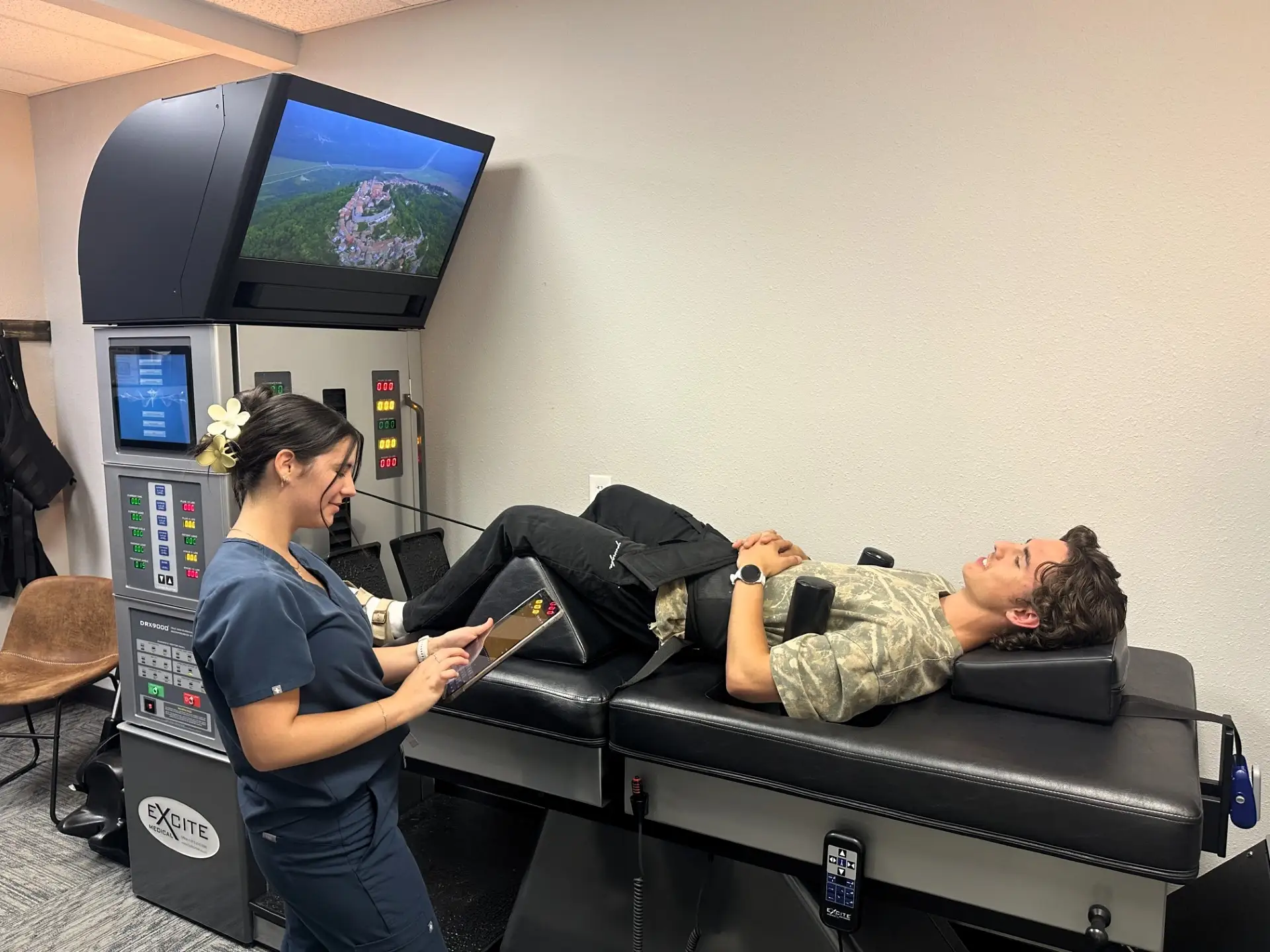 A technician assists a man lying on a medical treatment table, with a large screen displaying a scenic view above them.
