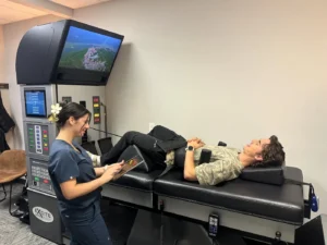 A female chiropractor monitors a male patient lying comfortably on a spinal decompression table, both smiling in a medical office.
