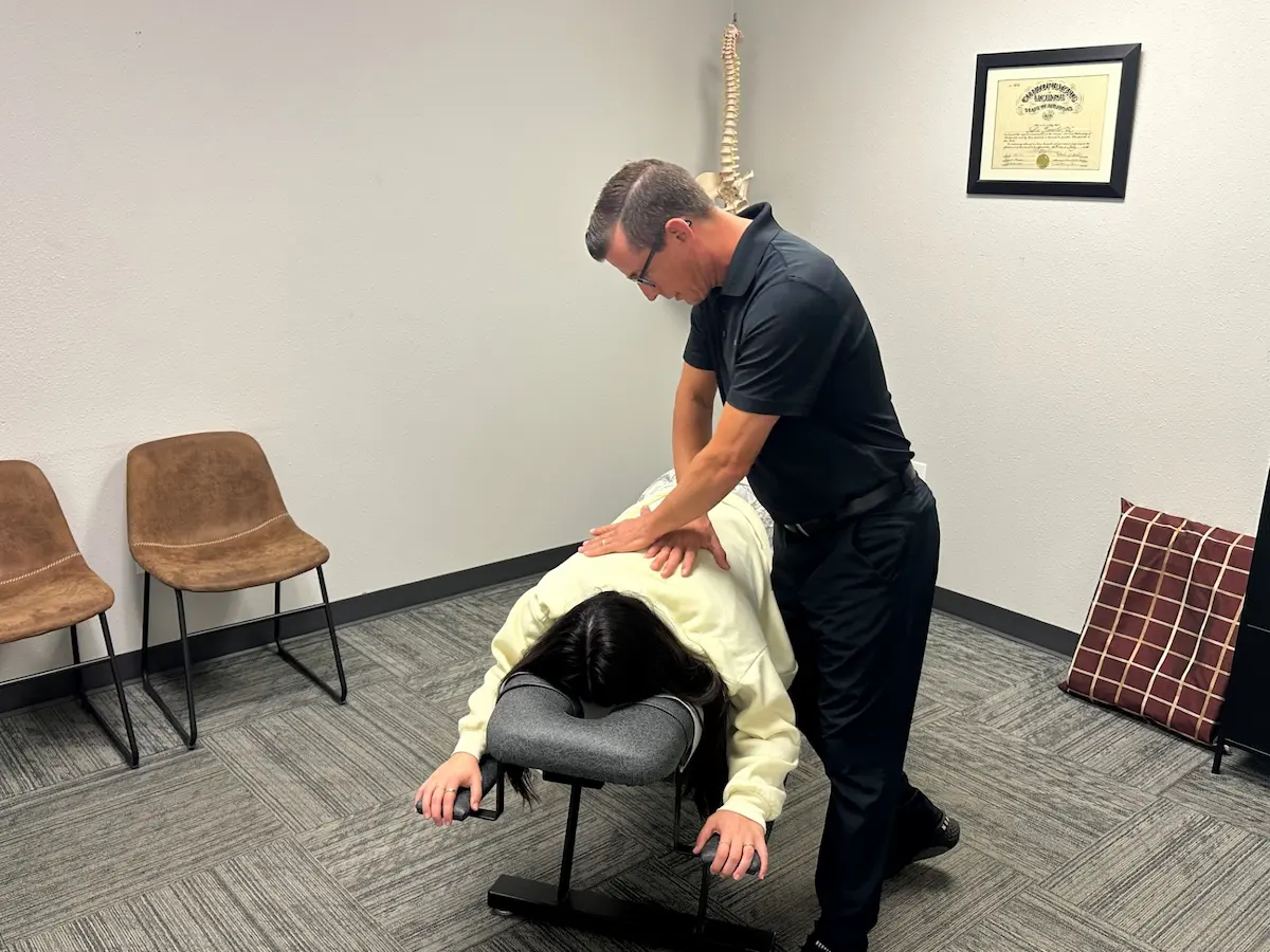 A chiropractor in a black shirt adjusts a patient lying face down on a chiropractic table. A spine model and framed certificate are on the wall.