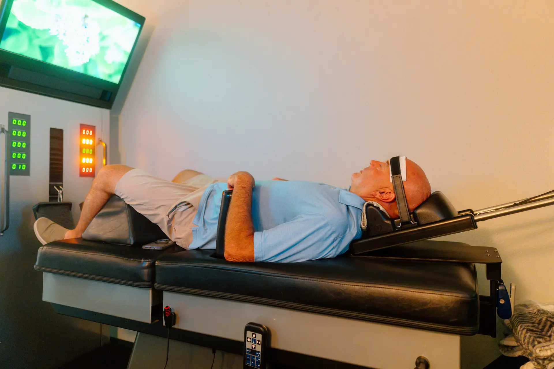 A man lies on his back on a therapy table with a head harness and knee support, facing a TV screen and control panels.