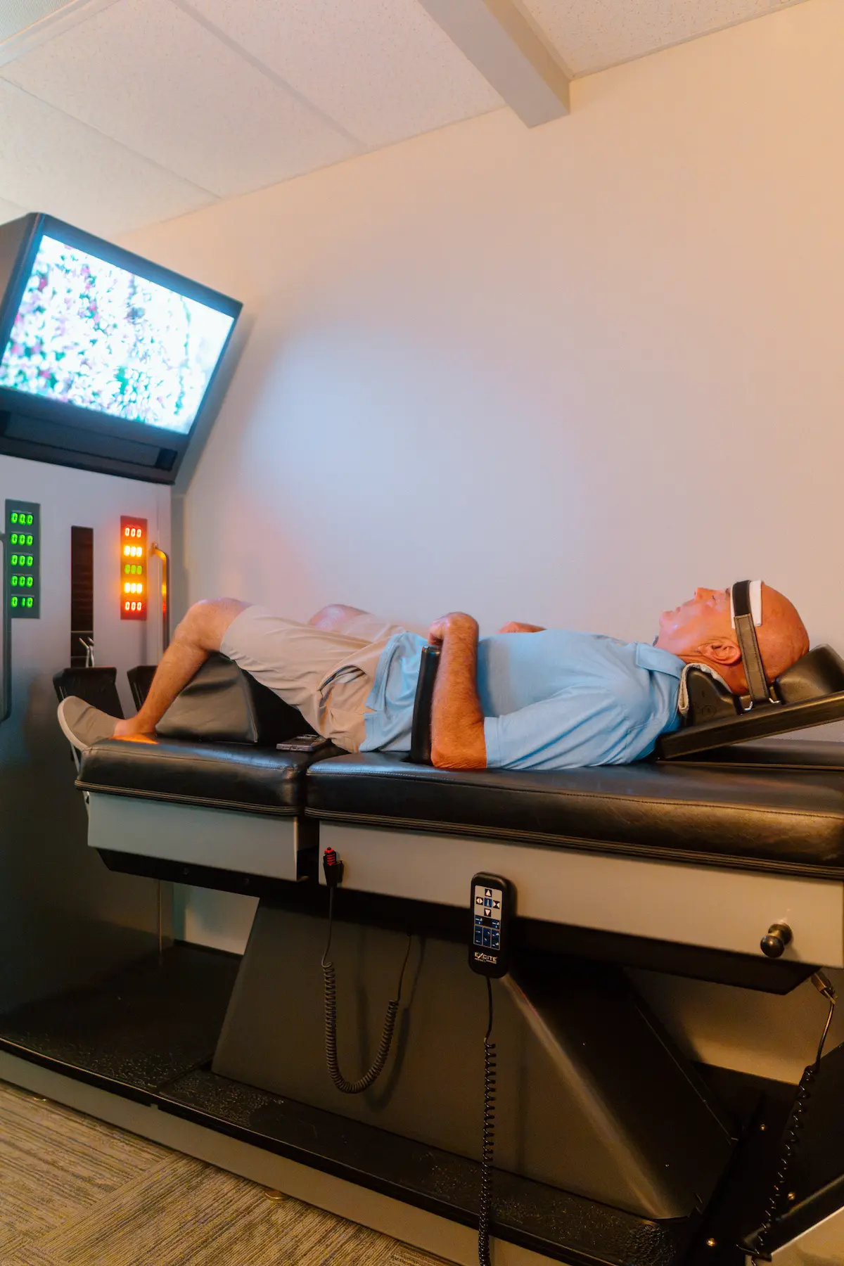 An older man lies on a therapy table with a head harness, looking at a monitor displaying abstract patterns, next to a control panel.