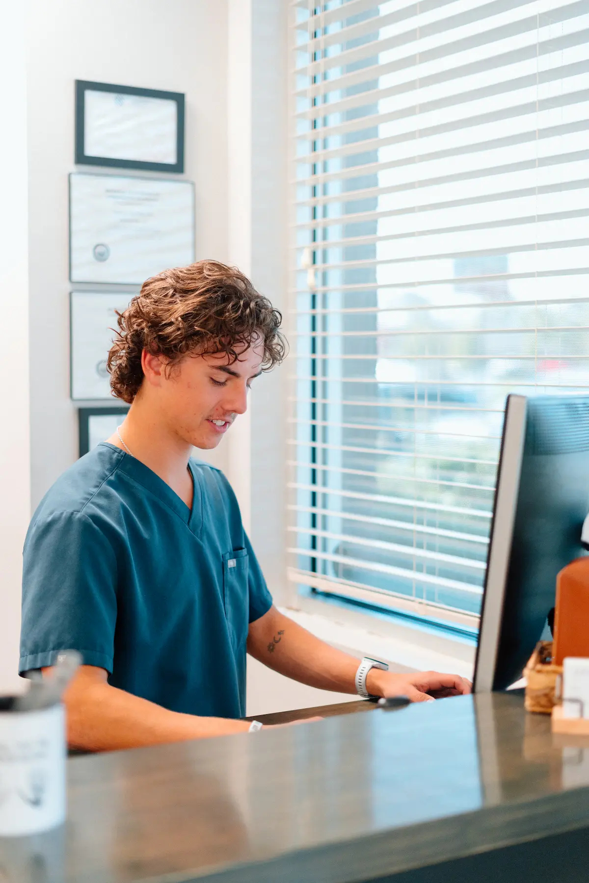 A young man in teal scrubs sits at a desk, looking at a computer with a slight smile.