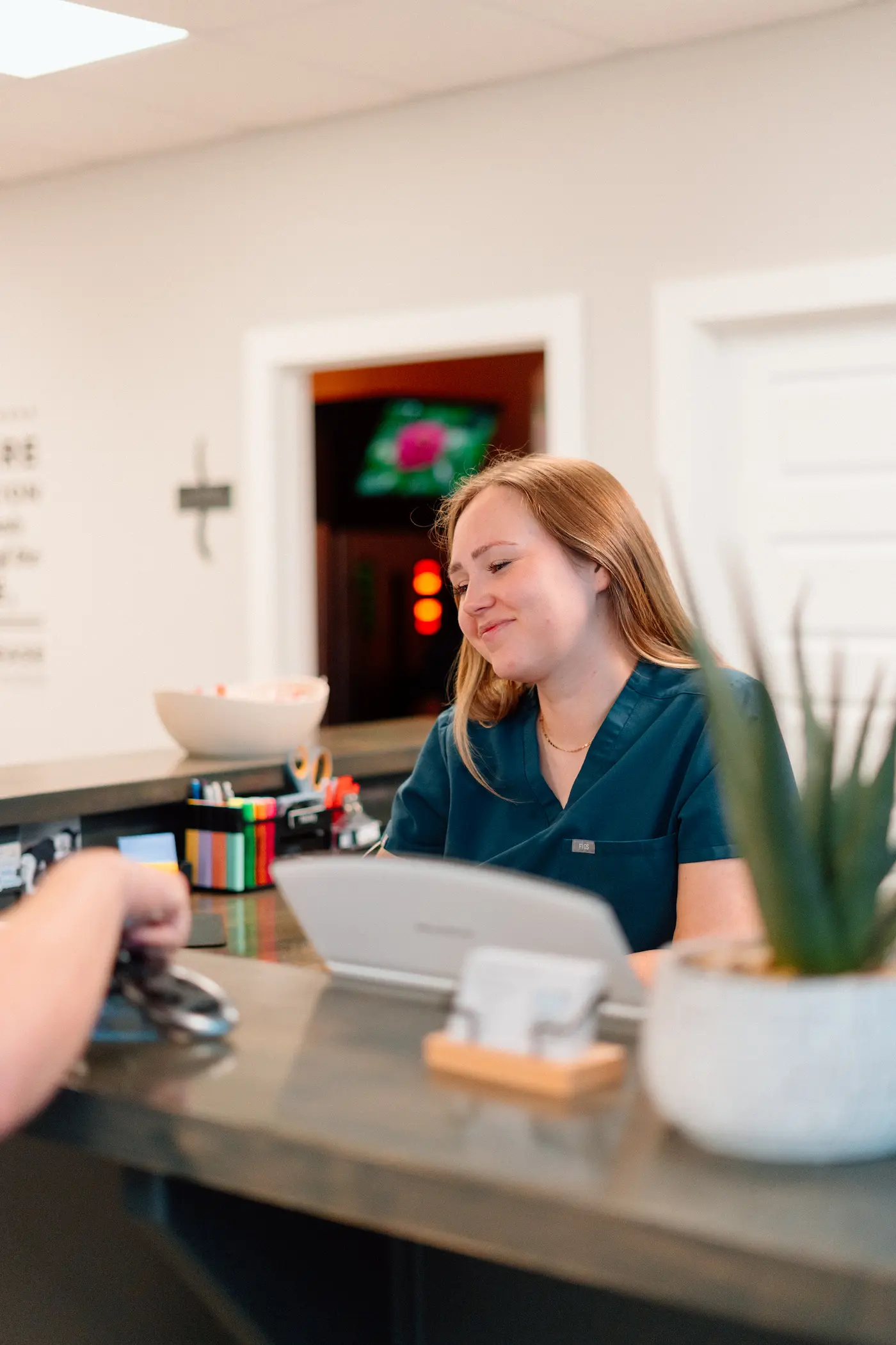 A smiling woman in teal scrubs works at a reception desk, looking at a tablet.