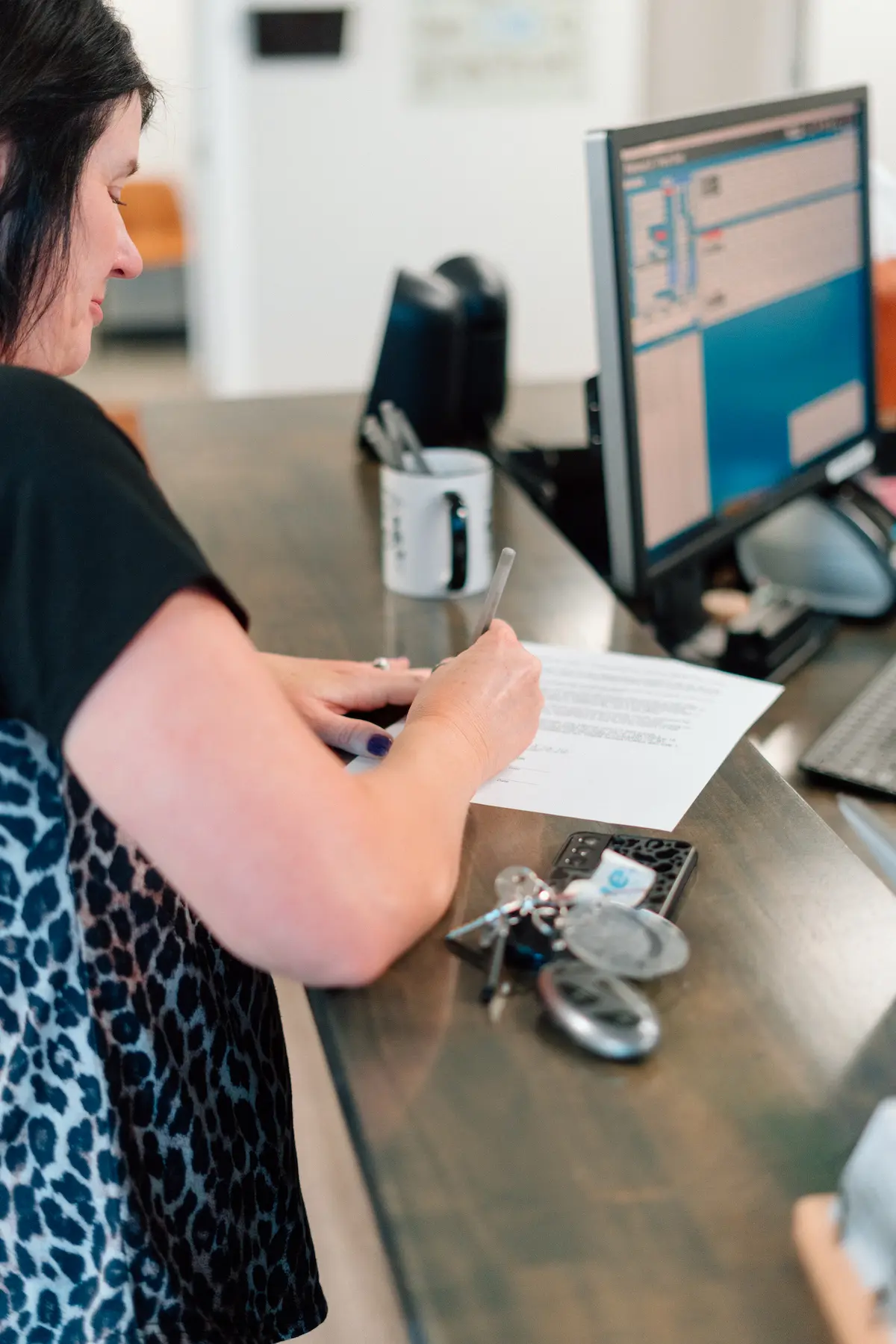 A woman in a black top and leopard print skirt writing on a document at a desk with a computer monitor, phone, and keys.