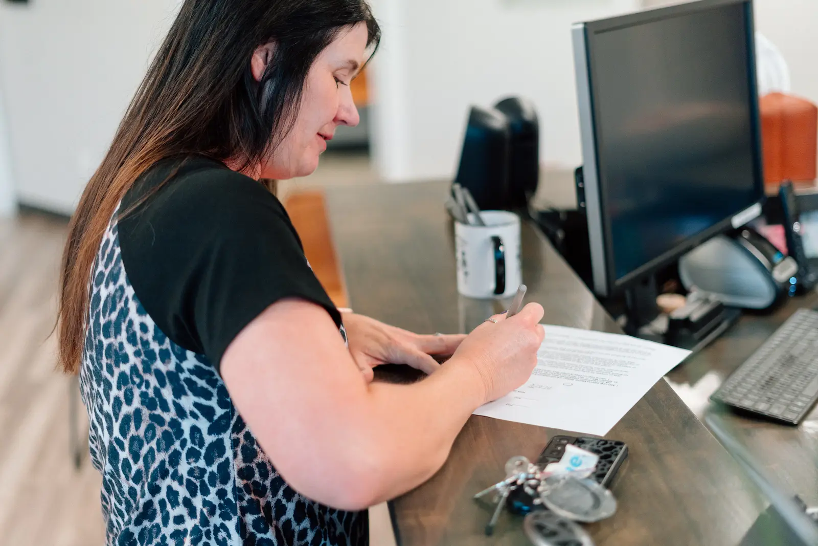 A woman with long dark hair, wearing a leopard print top, signs a document with a pen at a desk. Keys and a phone are next to the paper, with a computer monitor and mug in the background.