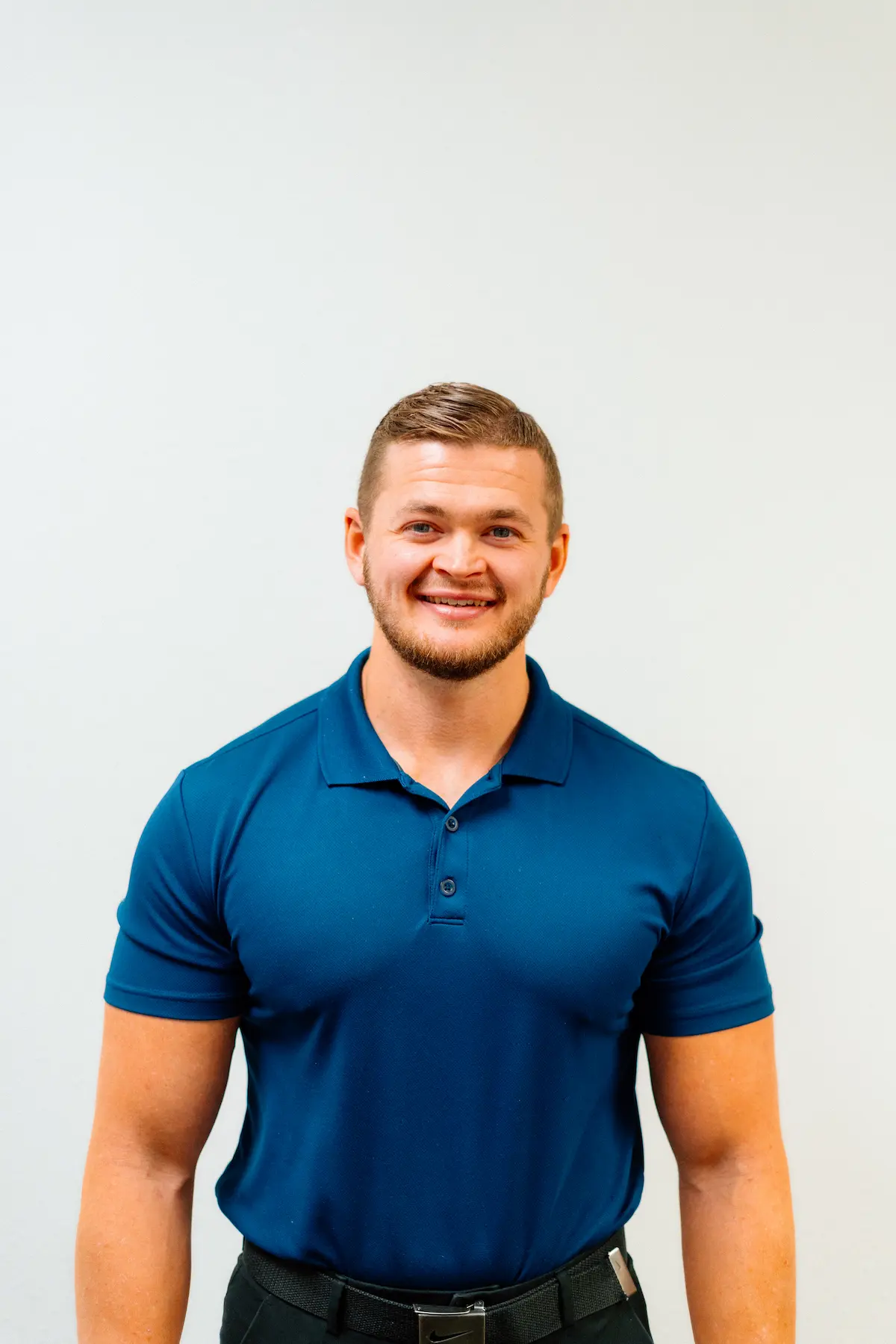 A man with short brown hair and a beard smiles at the camera, wearing a dark blue polo shirt against a light gray background.