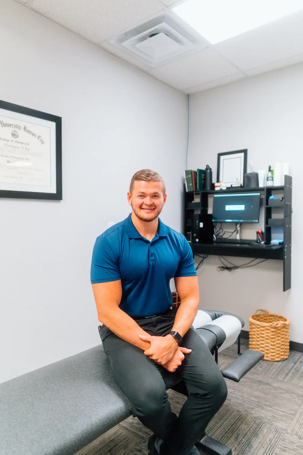 A smiling man in a blue polo shirt sits on a grey examination table in an office, with a diploma and computer desk in the background.
