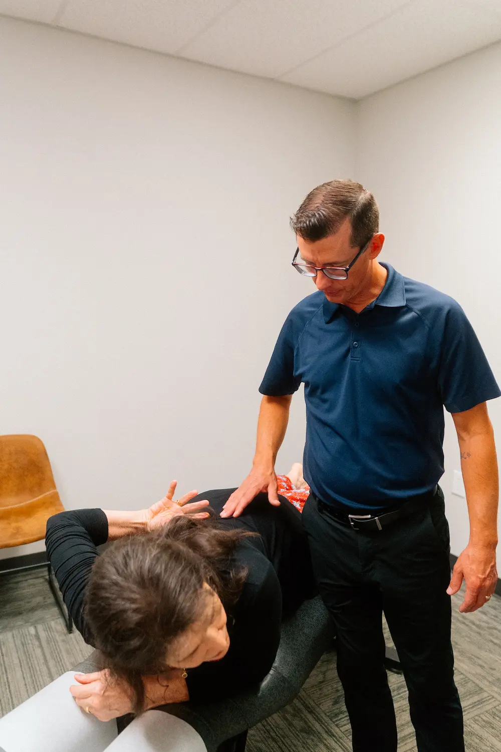 A man in a blue polo shirt adjusts a woman's upper back as she lies face down on an examination table.