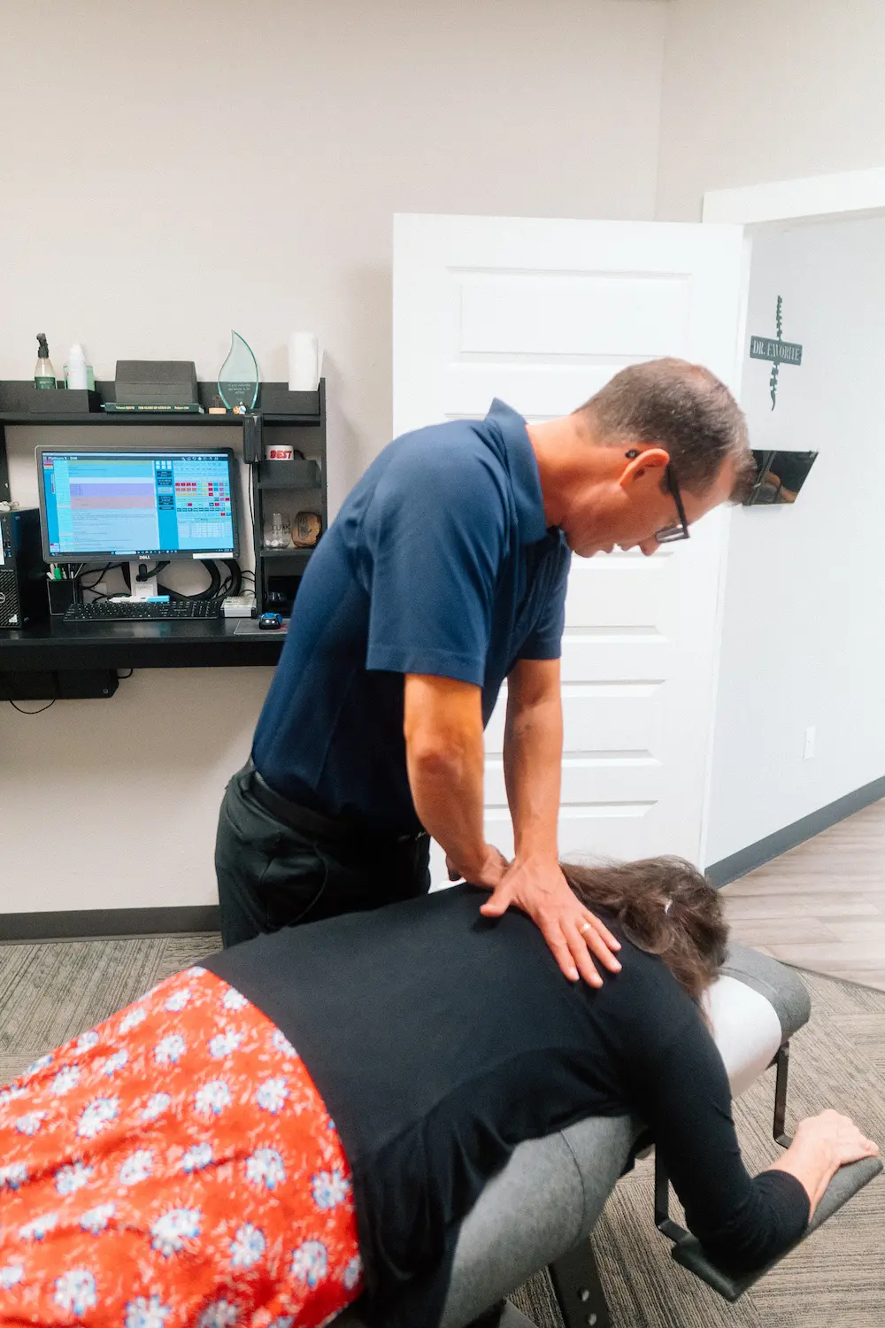 A male chiropractor adjusts a female patient's upper back on a grey treatment table in a clinic.