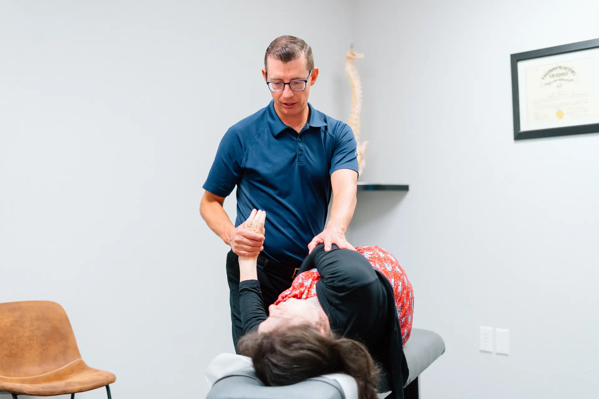 A male chiropractor adjusts a female patient's arm and shoulder on an examination table in a medical office.