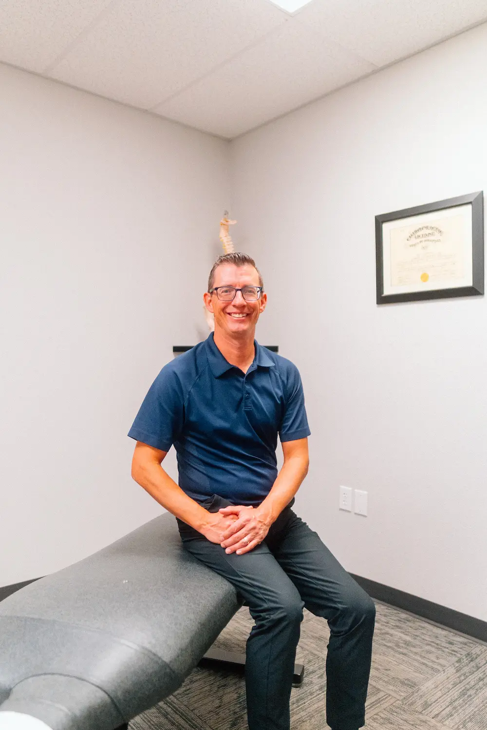A man in a navy polo shirt and glasses sits smiling on a grey examination table in a medical office.
