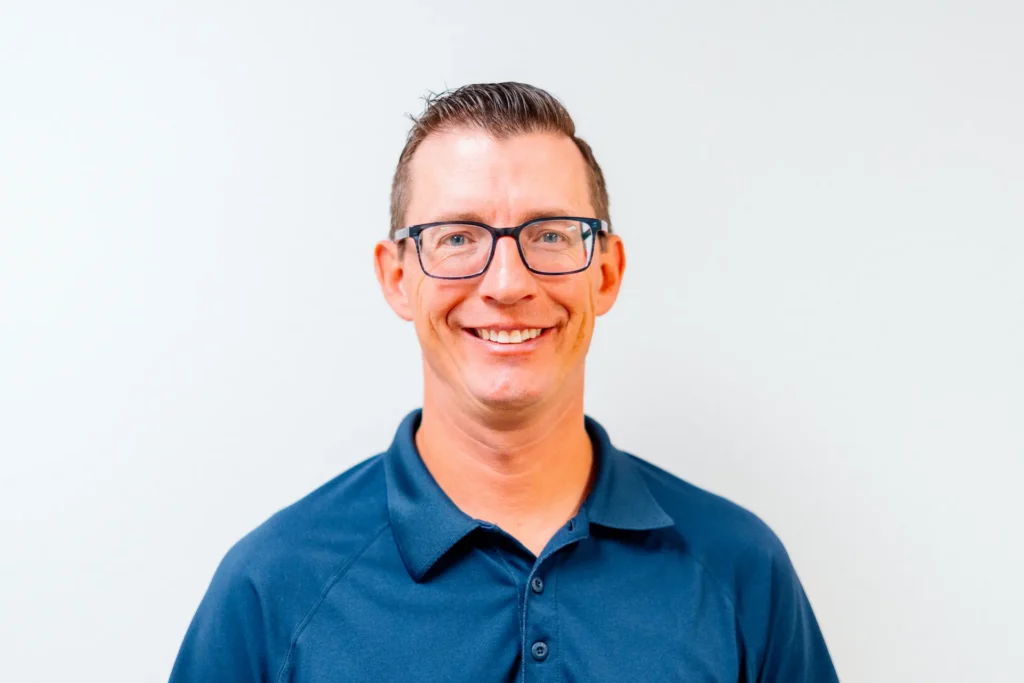 Headshot of a smiling man with dark hair and glasses wearing a blue polo shirt against a white background.