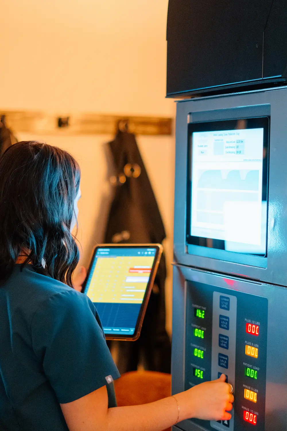 A person in teal scrubs operates a medical machine, viewing data on a tablet and the machine's digital displays and control panel.