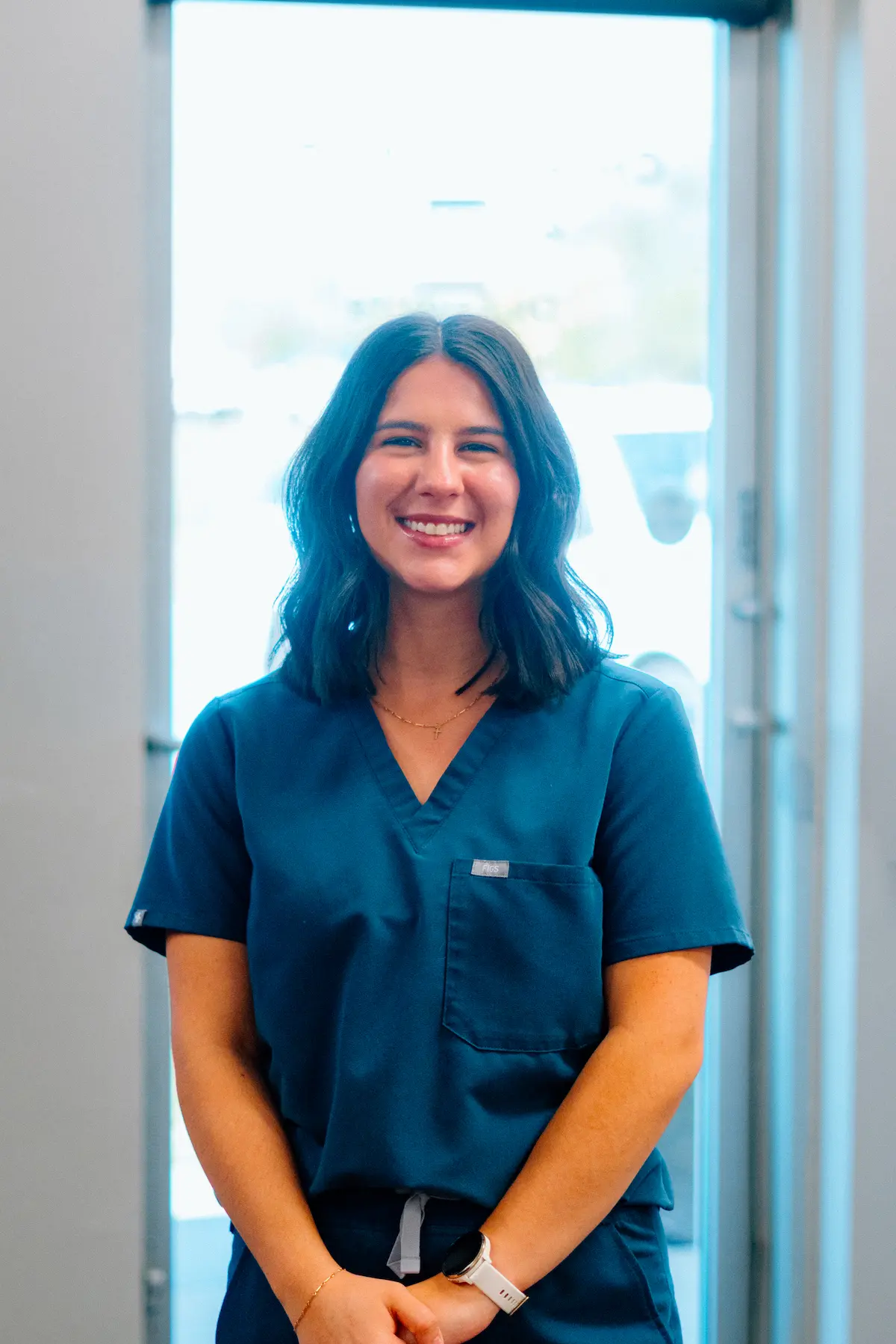 A smiling young woman with dark hair wearing teal scrubs, a gold cross necklace, and a white smartwatch, standing in front of a bright, blurred background.