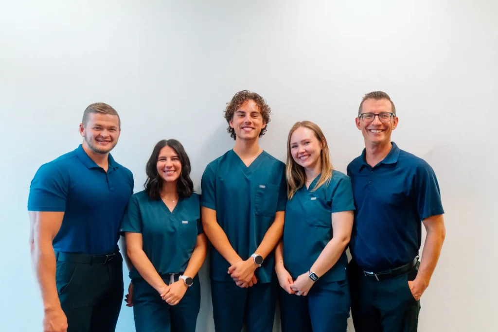 Five smiling individuals, three men and two women, stand in a line against a white wall. Two men wear blue polo shirts, and three others wear teal medical scrubs.