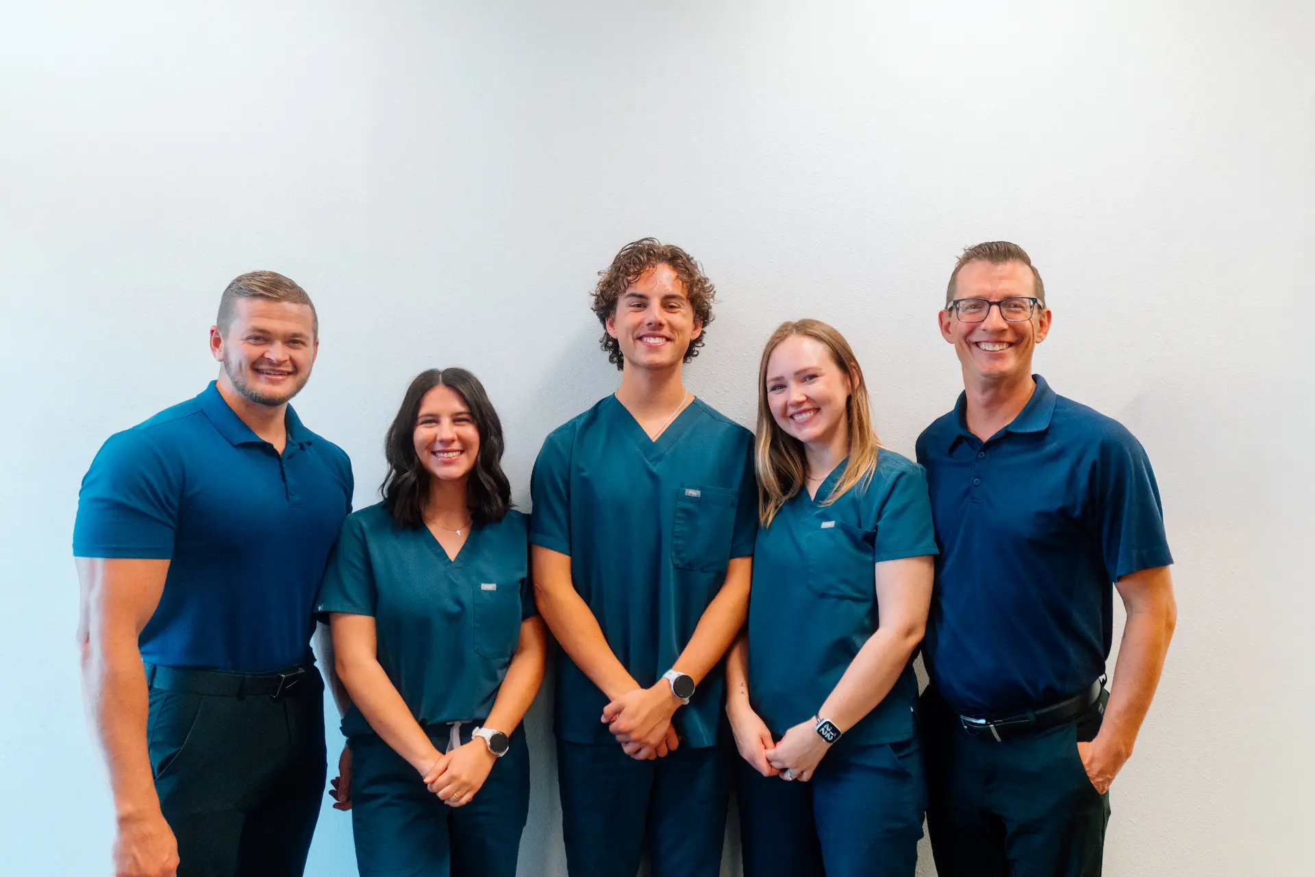 Five smiling individuals, three men and two women, stand in a line against a white wall. Two men wear blue polo shirts, and three others wear teal medical scrubs.