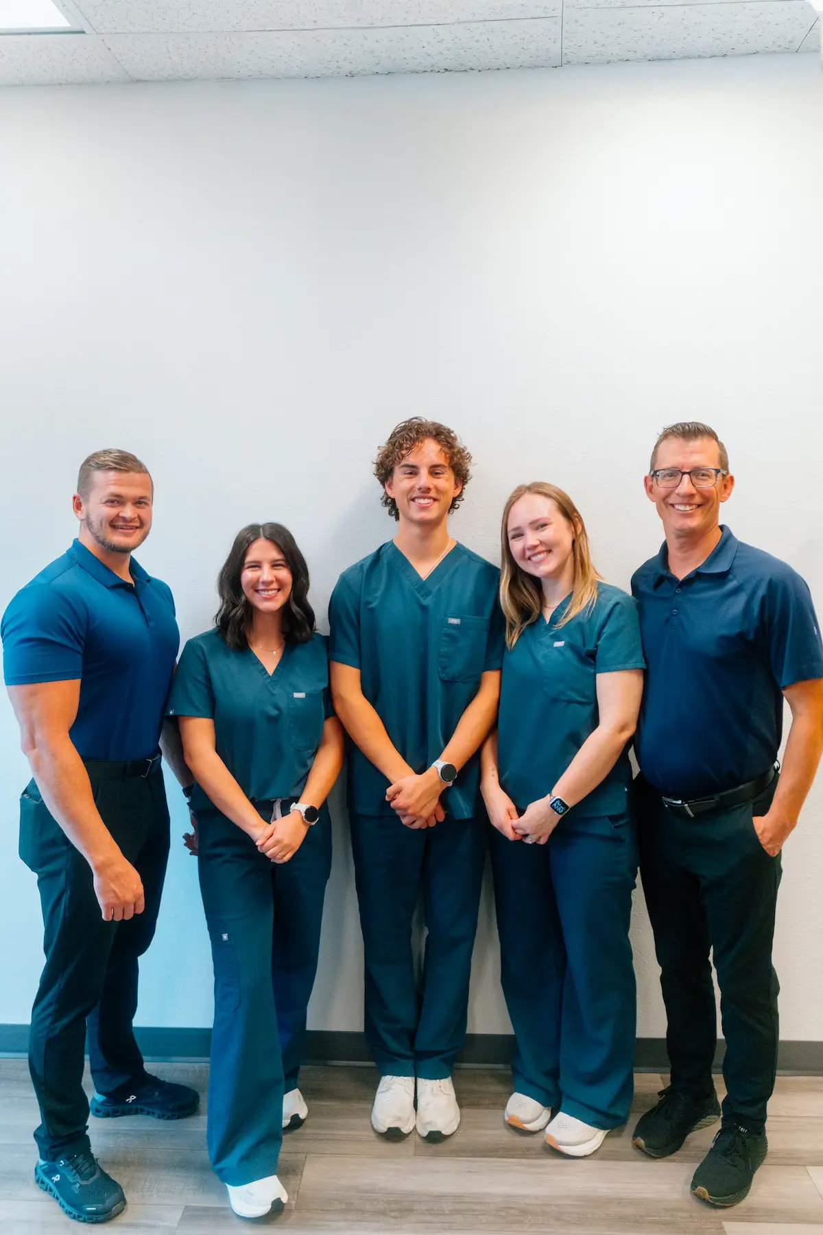 Group photo of five smiling people: three men and two women, some in dark blue polo shirts and others in dark teal scrubs, standing against a white wall.